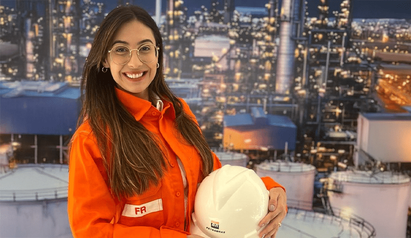 Jovem engenheira sorridente usando óculos e uniforme laranja da Petrobras, segurando capacete branco com logo da empresa, em frente a instalação industrial petrolífera iluminada