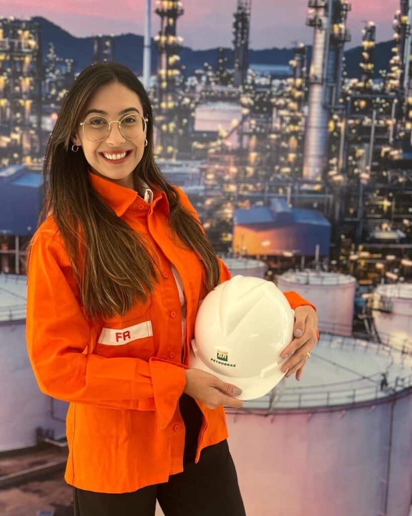 Jovem engenheira sorridente usando óculos e uniforme laranja da Petrobras, segurando capacete branco com logo da empresa, em frente a instalação industrial petrolífera iluminada.