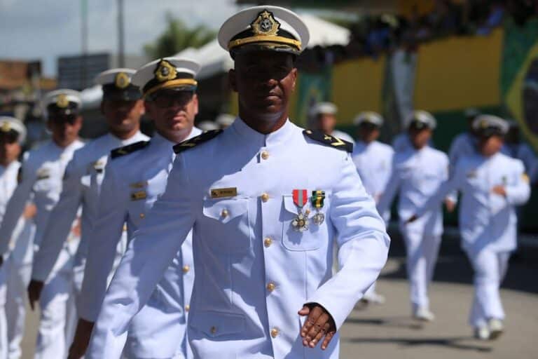 Oficial da Marinha do Brasil em uniforme branco durante cerimônia militar, com medalhas no peito e outros oficiais ao fundo em formação