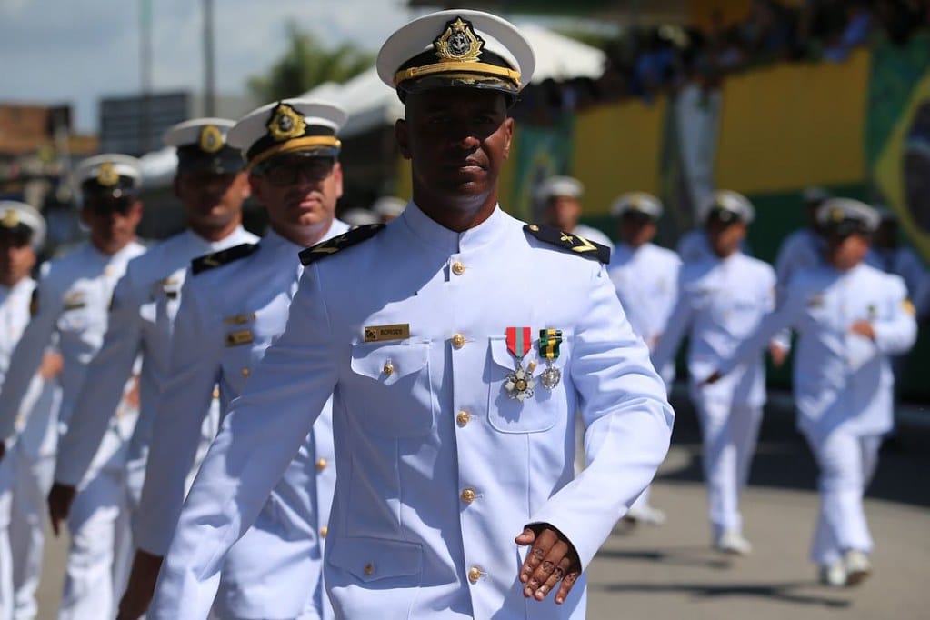 Oficial da Marinha do Brasil em uniforme branco durante cerimônia militar, com medalhas no peito e outros oficiais ao fundo em formação