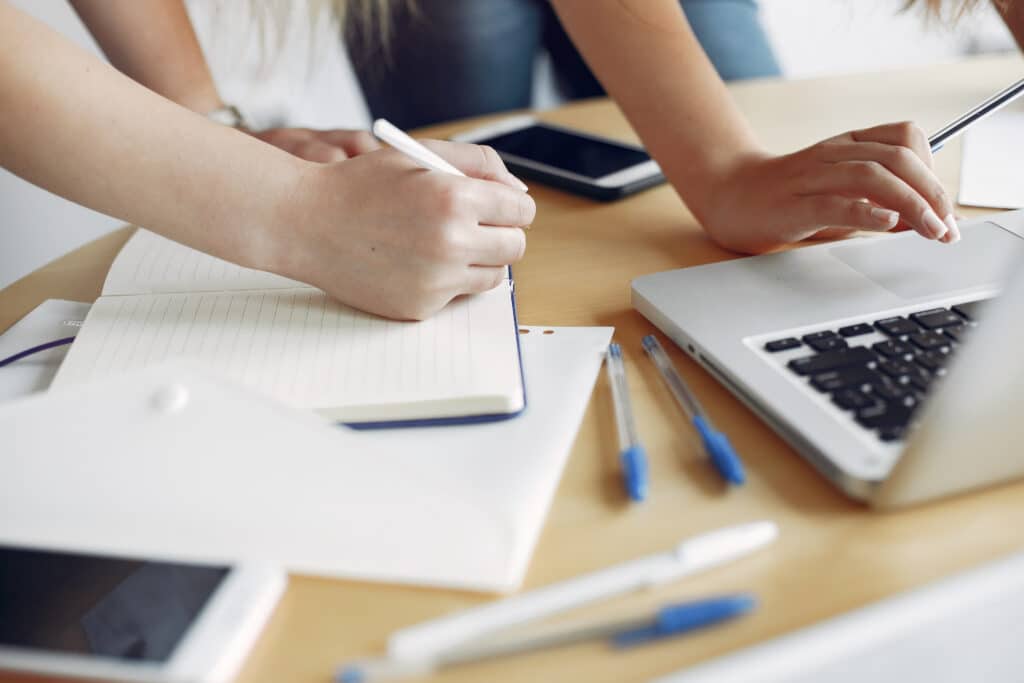 Pessoa estudando para concurso com caderno, caneta e laptop sobre mesa de madeira