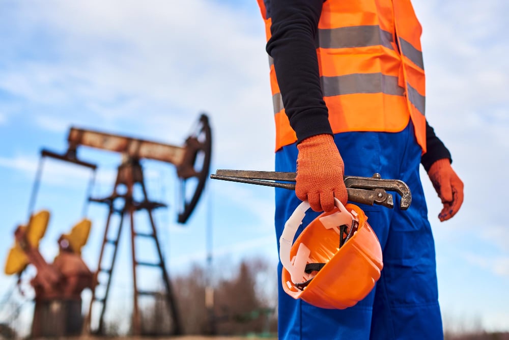 Profissional do setor petrolífero com equipamentos de proteção individual trabalhando em campo com bomba de extração de petróleo ao fundo
