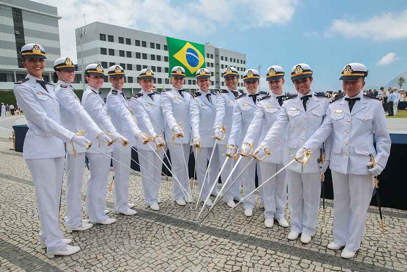 Grupo de oficiais da Marinha do Brasil em cerimônia de formatura com uniformes brancos e espadas cerimoniais em frente à bandeira do Brasil