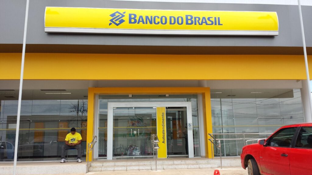 Aprovado no Banco do Brasil em frente a uma agência bancária com fachada amarela e logo do BB