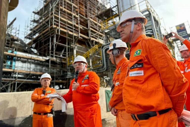 Profissionais da Petrobras em reunião operacional em instalação industrial com uniformes de segurança e capacetes durante atividade de campo
