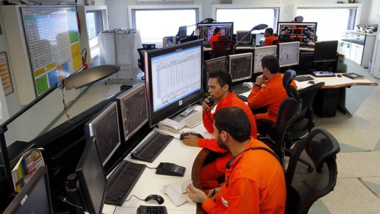Técnicos da Petrobras em sala de controle operacional monitorando equipamentos em plataforma offshore com múltiplos monitores e sistemas