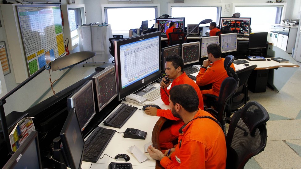 Técnicos da Petrobras em sala de controle operacional monitorando equipamentos em plataforma offshore com múltiplos monitores e sistemas