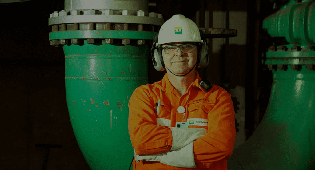 Profissional de segurança do trabalho da Petrobras com uniforme laranja, capacete e óculos de proteção em instalação industrial com tubulações