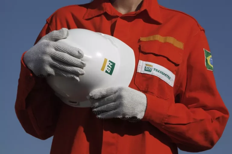 um profissional usando macacão vermelho da Transpetro, com a bandeira do Brasil na manga. A pessoa veste luvas de proteção cinza e segura um capacete de segurança branco com a marca BR. O fundo é azul e liso, destacando o uniforme e os equipamentos de proteção. O rosto não aparece na fotografia.